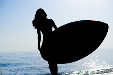 The dark silhouette of a slender girl surfer walks along the ocean and holds a board for  SUP water...