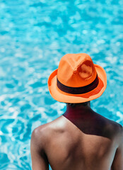 a boy of indian origin with his back to the blue water of a swimming pool. he wears a very striking orange hat. concept vacations and diversity. vertical format.