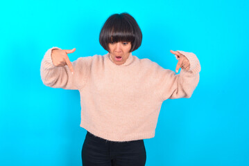 Amazed young brunette woman wearing pink knitted sweater over blue background points down with fore fingers, opens mouth being shocked. Advertisement concept.