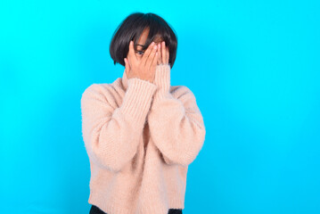 young brunette woman wearing pink knitted sweater over blue background covering face with hands and peering out with one eye between fingers. Scared from something or someone.