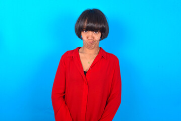 Fototapeta premium Offended dissatisfied young brunette woman with short hair wearing red shirt over blue background with moody displeased expression at camera being disappointed by something