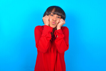 young brunette woman with short hair wearing red shirt over blue background with surprised expression keeps hands under chin keeps lips folded makes funny grimace