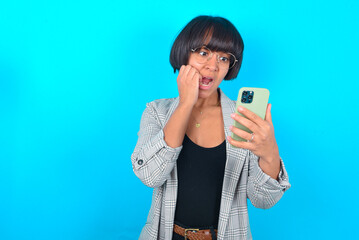 Fototapeta premium Portrait of pretty frightened Young businesswoman with bob haircut wearing blazer against blue wall chatting biting nails after reading some scary news on her smartphone.