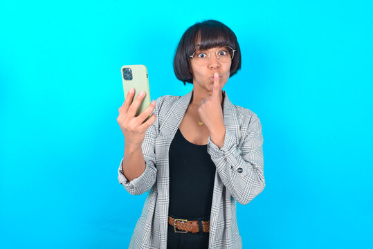 Young businesswoman with bob haircut wearing blazer against blue wall holding modern gadget ask not tell secrets