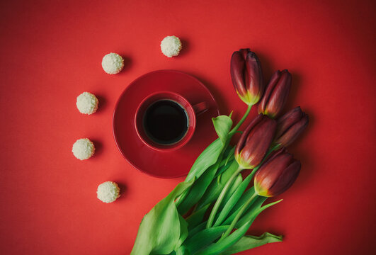 A cup of coffee in a red cup on a red background with red tulips and white round candies next to it