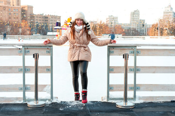 Beautiful lovely young adult woman brunet hair warm winter jackets stands near ice skate rink...
