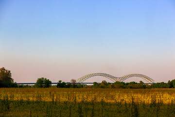 Fototapeta na wymiar The Hernando de Soto Bridge in West Memphis, Arkansas crossing the Mississippi river to Memphis Tennessee. With and open field of yellow flowers, blue sky and trees. Photographer Derek Broussard