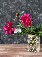 A bouquet of peonies with a greeting card in a vintage handmade vase on a wooden table and black concrete background with copy space. Rustic still life with summer flowers. Pink peony.