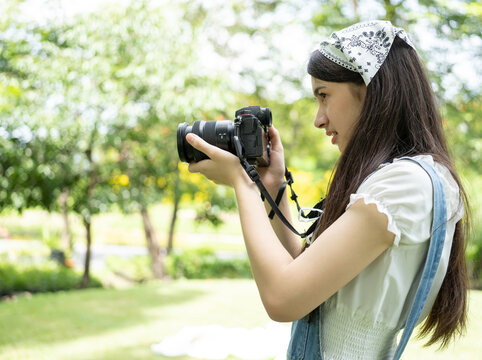 Beautiful woman holds digital camera and taking photo blurry background. Behind the scene, young Asian photographer standing in green park taking pictures as recreation. Tourist photographing concept.