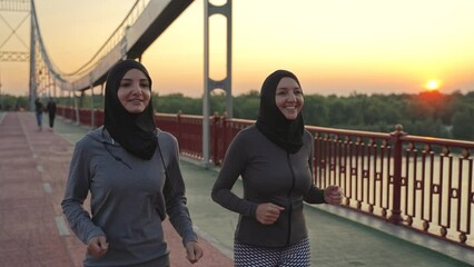 Smiling fit females in hijabs and sportswear jogging on footbridge across city river at dawn. Sporty arab runners enjoying joint morning workout, active healthy urban sports lifestyle concept