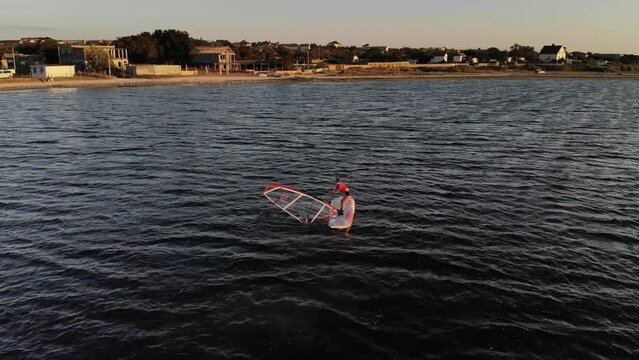 A novice windsurfer girl is learning to steer a windsurfing board with a sail on a lake at sunset