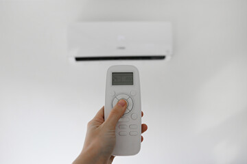 Close up shot of a woman's hand holding the remote control of newly installed white air conditioner. AC hanging on the wall working in a cooling mode set to 24 degrees celsius. Background, copy space.