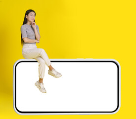 Fototapeta na wymiar Thoughtful asian girl sit on huge smartphone with white screen wearing casual white jeans isolated on yellow background. Mock up product placement. Mobile App Advertisement. Copy space