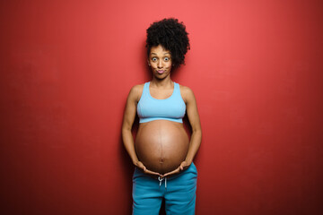 Fun portrait of surprised healthy expressive black pregnant woman on fitness sportswear against red background. - 510600733