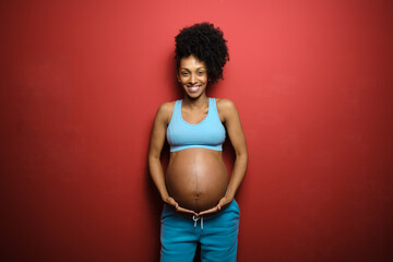 Portrait of blissful black pregnant woman on fitness sportswear against red background. - 510600734