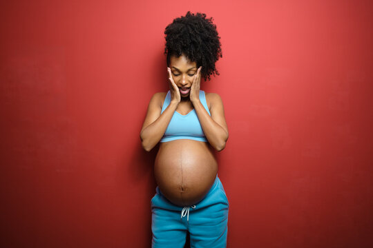 Fun portrait of surprised healthy expressive black pregnant woman on fitness sportswear against red background.