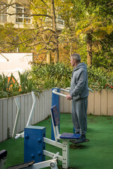 An elderly gray-haired man pensioner in a gray suit is training on simulators on a sports field on the street. The concept of an active healthy lifestyle of elderly pensioners.