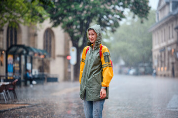 Fototapeta premium Young woman in green yellow raincoat stands happily on street under the rain