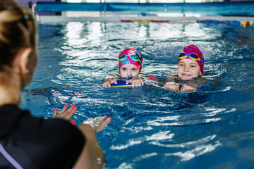 Children learn to swim with board in pool under guidance of coach. Active kids are playing in water.