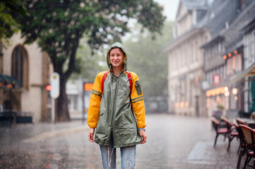 Young woman in green yellow raincoat stands happily on street under the rain