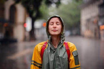 Young woman in green yellow raincoat stands happily on street under the rain