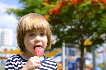 Portrait of a cute boy on the background of a bright playground. The child eats a heart-shaped...