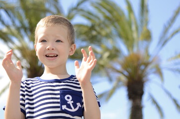 Positive emotional boy blond, 5 years old in a sailor's T-shirt against the backdrop of palm trees. Concept: traveling with children, family resort, summer, sailing.