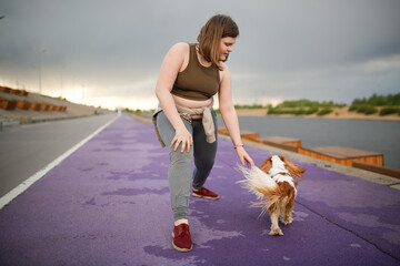 European teenage girl overweight on jog on treadmill along embankment of city with dog , overweight...