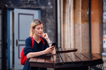 Fototapeta premium Young blonde woman in autumn-spring outwear and red backpack stands near high wooden table