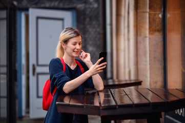 Young blonde woman in autumn-spring outwear and red backpack stands near high wooden table