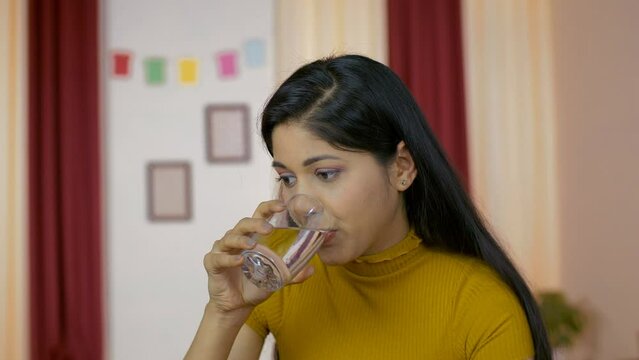 Thirsty young woman holding and drinking a glass of fresh and pure water. Medium shot of an Indian girl following a good lifestyle/weight loss plan and drinking lots of water - health care concept