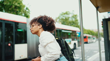 Young woman in glasses looking at a smartphone while standing at a bus stop. Positive woman using...