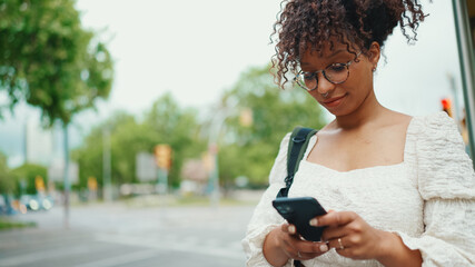 Young woman in glasses looking at a smartphone while standing at a bus stop. Positive woman using mobile phone outdoors in urban background.