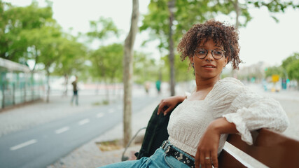 Young woman in glasses sits on a bench smiling and looking around. Happy girl looks around sitting on a bench - Powered by Adobe