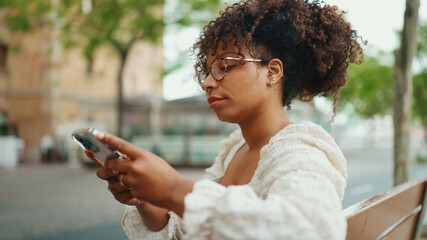 Closeup portrait of a young woman in glasses sits on a bench smiling and sends a voice message. Girl using mobile phone outdoors in urban background.