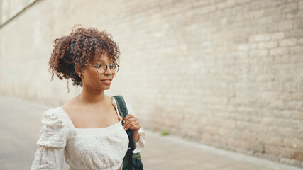 Young woman in glasses walks down the street along a brick wall. Girl goes on the street in urban background