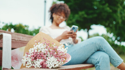 Young woman sits in the park on a bench with a bouquet of flowers with a smartphone in her hands. Positive woman using mobile phone outdoors in urban background.