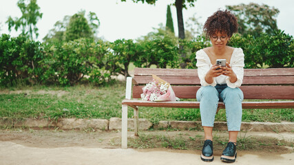 Young woman sits in the park on a bench with a bouquet of flowers with a smartphone in her hands. Positive woman using mobile phone outdoors in urban background.