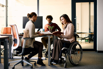 Fototapeta na wymiar Happy businesswoman in wheelchair going through reports with her colleague in office.