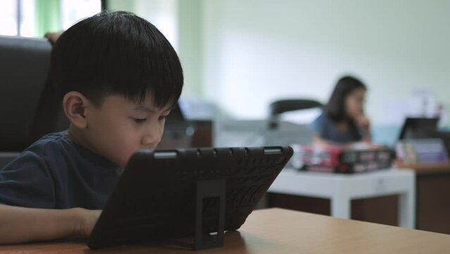 Asian boy addictive watching tablet while his mother working on the computer in the background.