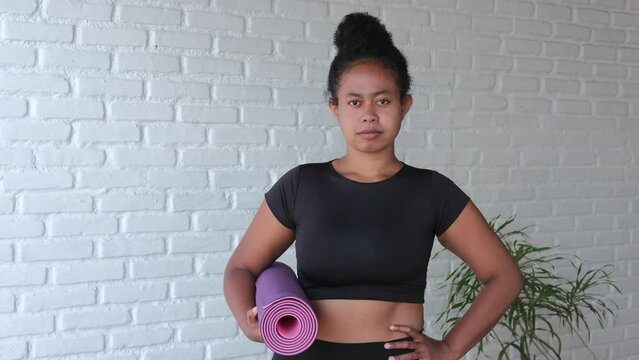 In a bright room, daylight falls on a standing in sports black clothes holding a rolled-up purple fitness mat with her right hand. I put my other hand on my thigh for convenience. zambo model