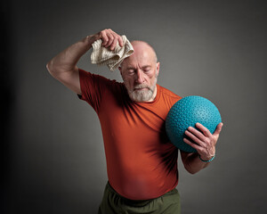 senior man (in late 60s) is exercising with a heavy slam or medicine  ball and wiping sweat with a towel, activity and fitness over 60 concept