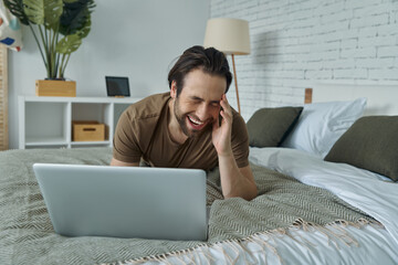 Cheerful young man using laptop and laughing while lying on bed at home