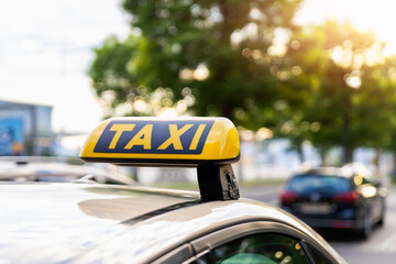 Close-up detail yellow taxi symbol on cars roof stand waiting at parking of airport terminal or railway station against park warm evening bokeh sunlight. Urban street transportation comfort service - obrazy, fototapety, plakaty
