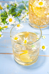 Herbal organic cold drink. Infused flower water. Iced daisy Chamomile tea or lemonade with lemon slice and ice cubes, in two different glasses, blue wooden background copy space