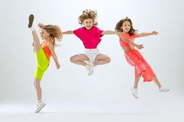 Three little girls, kids in bright colorful clothes dancing, posing isolated on white studio background. Concept of music, fashion, art, childhood, hobby