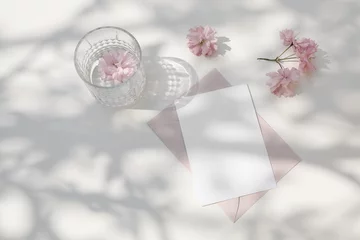 Foto op Canvas Wedding sationery. Pink Japanese cherry tree, sakura blossoms and glass of water, drink in sunlight. Greeting card, invitation mockup. Pink envelopes. White dappled table background, shadows. Flat lay © tabitazn