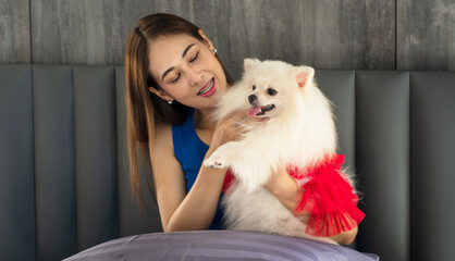 Asian girl lounging on the bed in the bedroom at home hugging a little white dog, a loving pet, happy playing with a furry puppy.