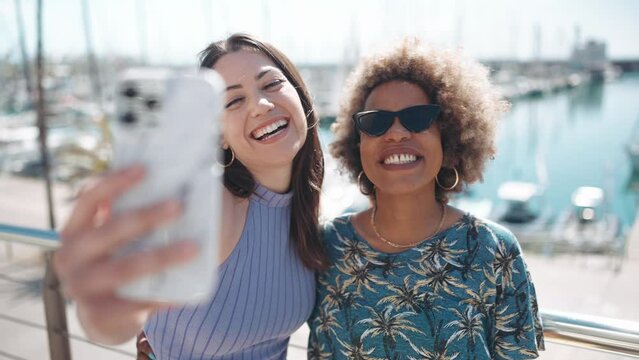 Two smiling multiethnic women making selfie on phone on the embankment near the yachts  