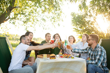 Group of young friends enjoying a picnic sitting outdoors fresh air spending weekend sunny day drinking beverage clinking cups, soft focus